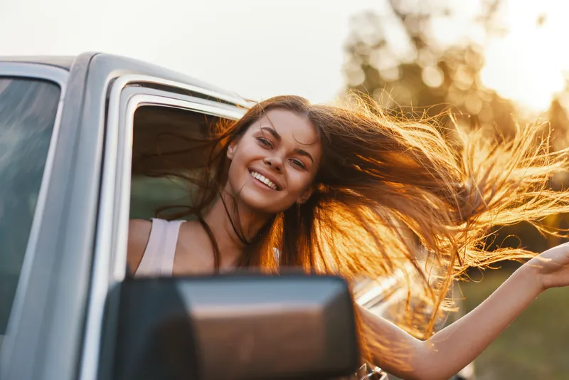 Young woman with long hair smiling while traveling in a car, capturing the essence of freedom and adventure during a sunny day in nature.