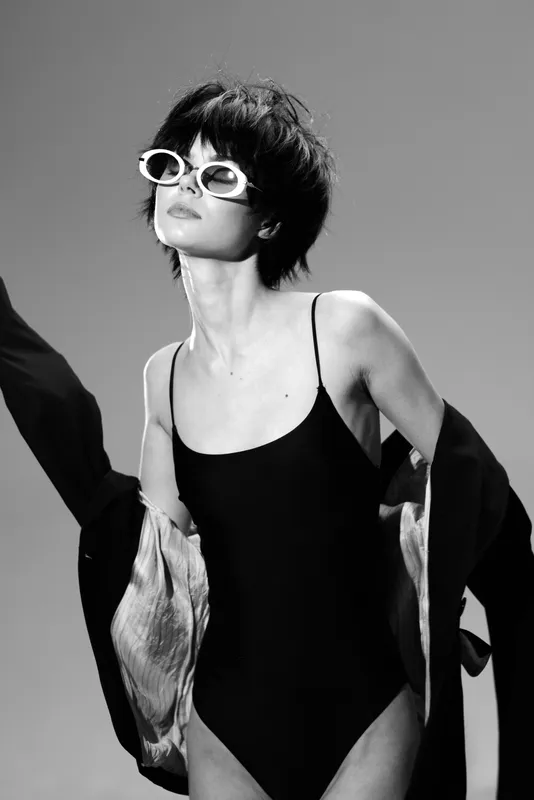 Black and white portrait of stylish woman in sunglasses wearing a swimsuit and open jacket, posing confidently with a retro vibe.