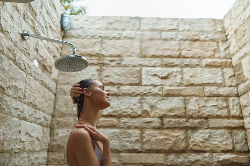 woman showering outdoors under rainfall showerhead on stone wall background, natural light, peaceful moment, wet skin, spa setting, relaxation