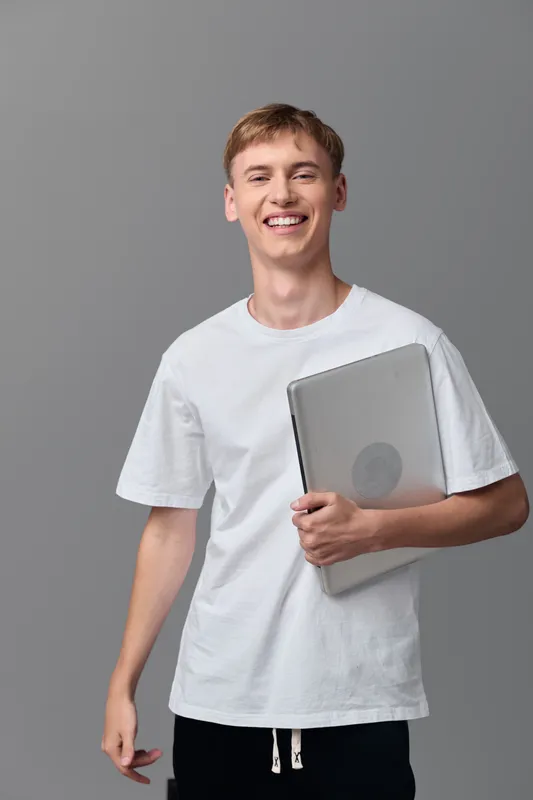 Smiling young man in a white t shirt holding a laptop, studio backdrop, casual learning and technology concept for education, youth and workplace use