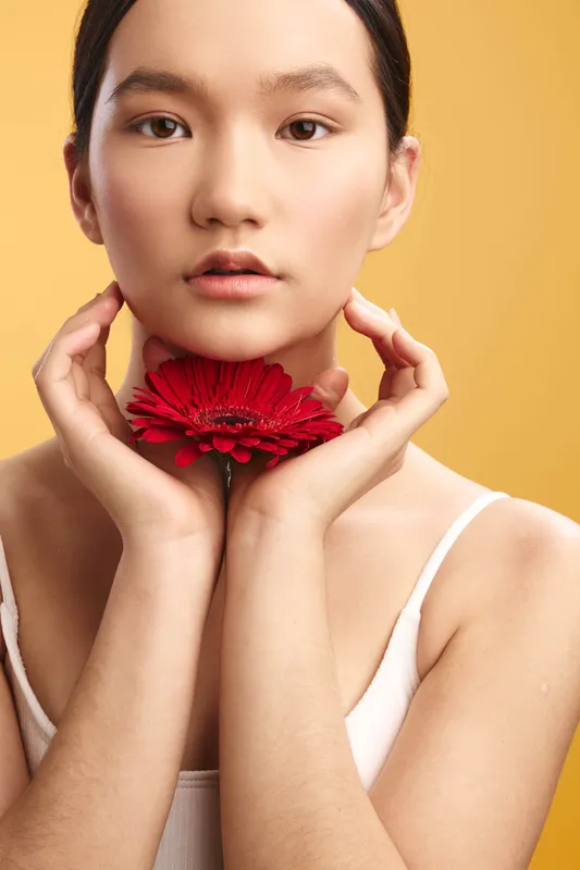 asian woman holds a red flower near her chin in a soft studio portrait, calm expression, warm light, minimalist background, gentle beauty, quiet elegance, focus on detail and serenity.