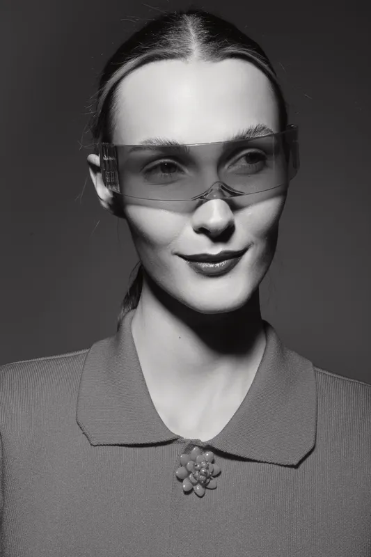 Young white woman with pale skin wearing transparent glasses and a vintage collar brooch looks aside on dark background.