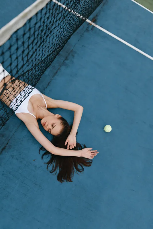 Young woman resting on a tennis court in a sporty white outfit, embodying relaxation and serenity, surrounded by blue tones with a tennis ball nearby