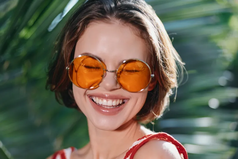 A cheerful woman wearing orange sunglasses smiles outdoors, sunlight framing her face. She exudes warmth and confidence, capturing a vibrant, carefree mood suitable for lifestyle and summer themes.