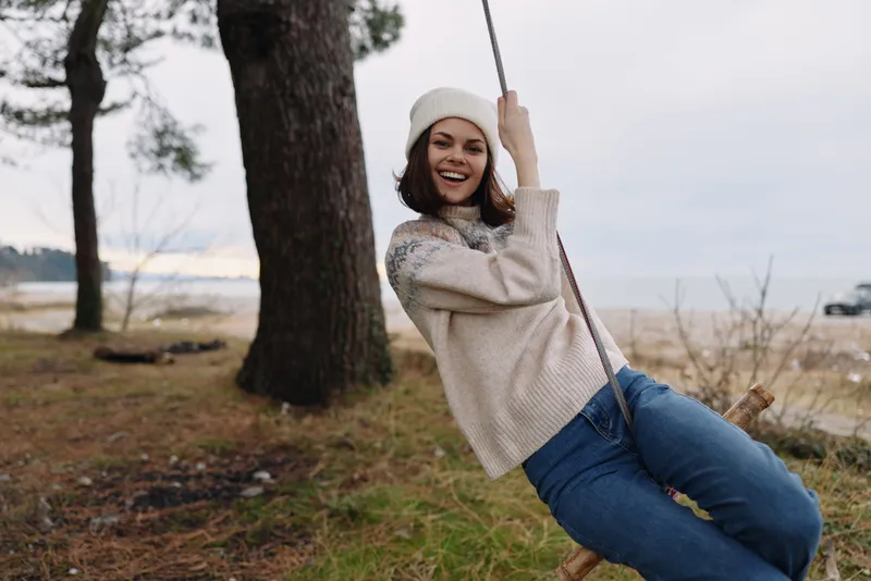 Woman on a rope swing outdoors, smiling in a cozy sweater and beanie, surrounded by trees in a quiet park, enjoying a carefree moment in nature.