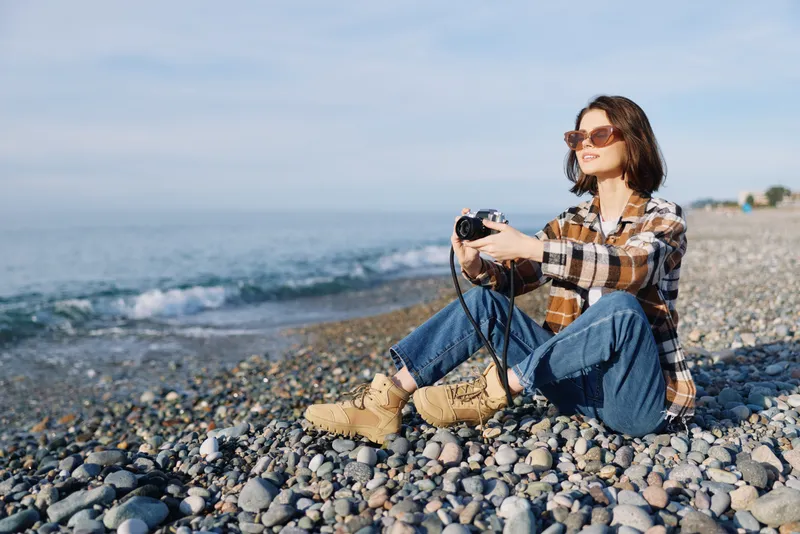 Photography scene of a woman on a pebble beach with a camera, wearing a plaid shirt and jeans, enjoying outdoor photography and sea breeze while capturing coastal scenery.