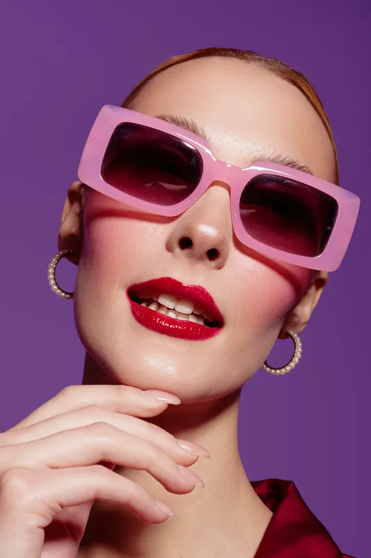Close-up of fashionable woman wearing pink sunglasses and red lipstick with hoop earrings on purple background. Concept of style, beauty, and modern fashion accessories.