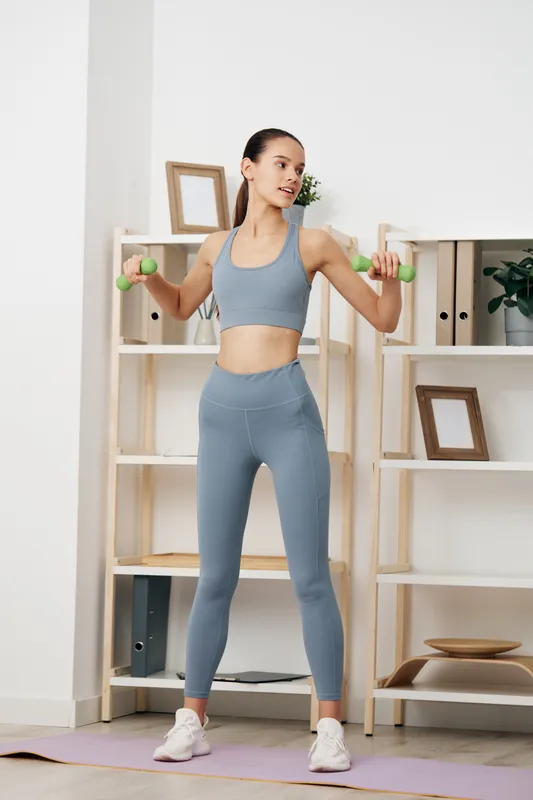 Fitness woman in gray athletic set lifting dumbbells during a shoulder workout in a bright home gym with shelves and a plant.