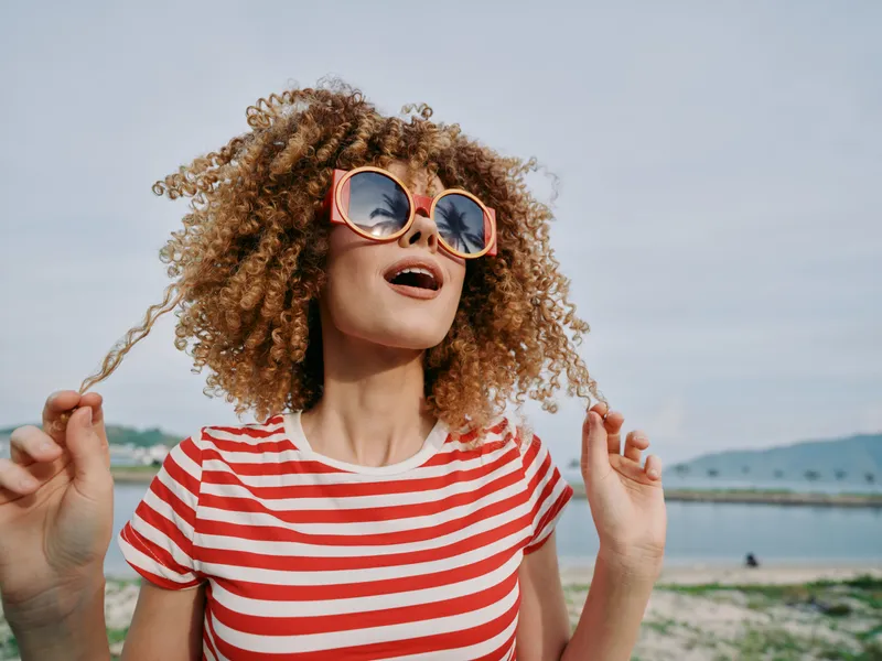 Portrait of a joyful woman with curly hair wearing pink sunglasses and a red white striped tee, standing outdoors near water under a clear sky.