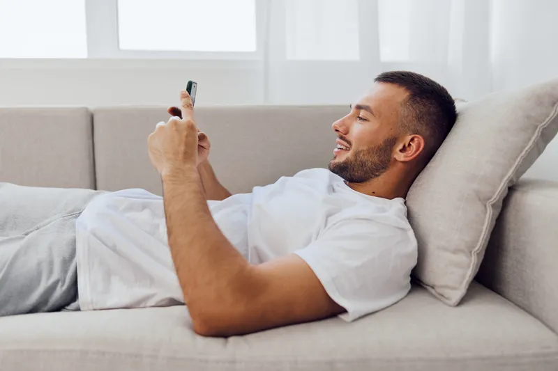 Relaxed man engaging with technology while lounging on a couch, reflecting themes of digital detox and work-life blur. A candid moment of solitude and self-care.