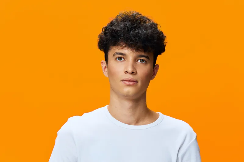 Young man with curly dark hair wearing a white t-shirt poses confidently against a vibrant orange background in a studio portrait setting.