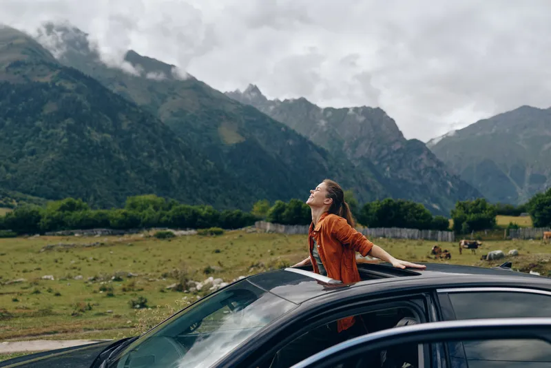 Person leaning out of a car window with arms outstretched, enjoying a scenic mountain landscape under a cloudy sky, conveying freedom and adventure for a bold travel mood