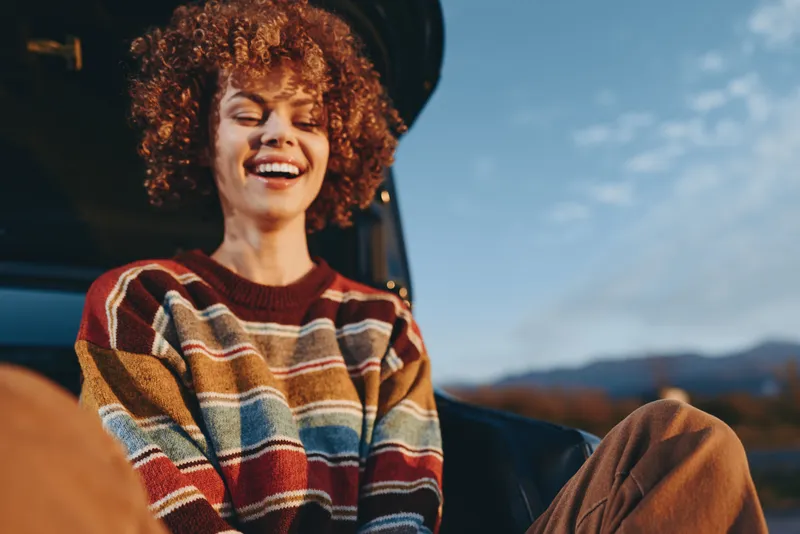 Woman with curly hair smiles in a striped sweater while relaxing in an open car outdoors, with mountain landscape blurred behind. Casual lifestyle travel moment under evening light.