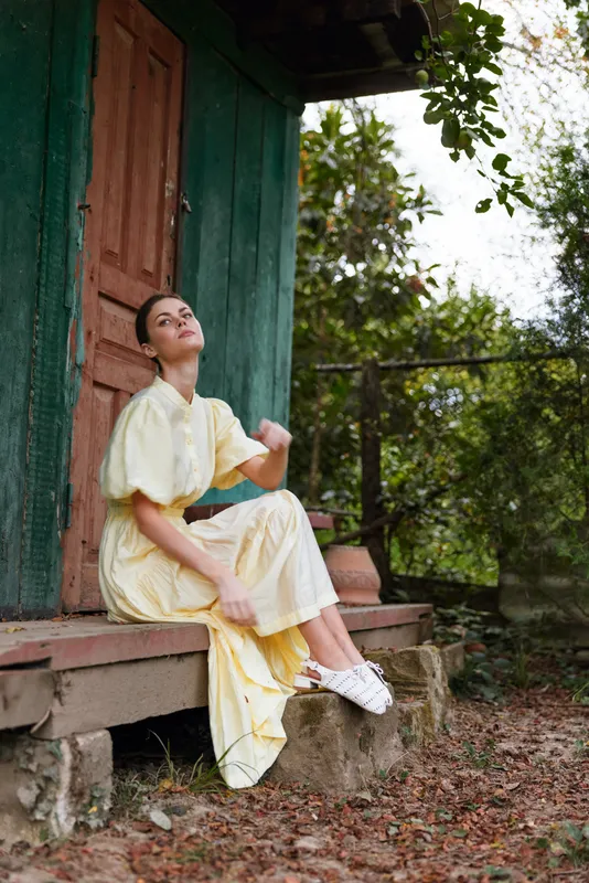 woman sitting on rustic porch in yellow dress and sneakers looking up thoughtful countryside setting with nature background authentic inclusive portrait calm