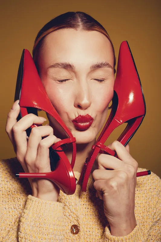 Young woman with closed eyes holds red high heels near face, wearing yellow sweater. Concept of fashion, style, beauty, and footwear with vibrant colors and joyful mood in studio.