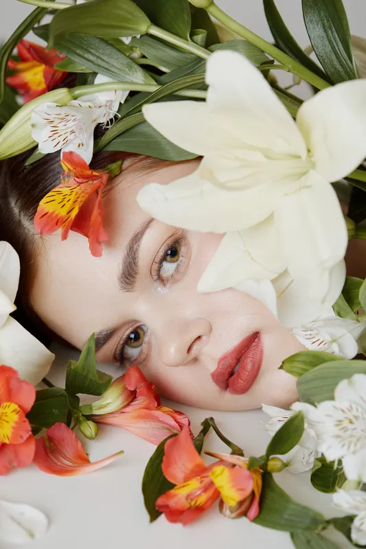 Young model with a serene expression surrounded by vibrant flowers in a soft, neutral backdrop, showcasing beauty and nature
