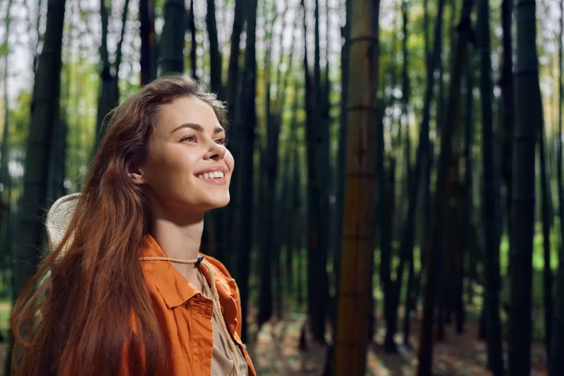 Woman, smile, bamboo, forest, portrait, nature, outdoors - Young woman with long hair and orange jacket smiling and looking up among tall bamboo trees in a peaceful green woodland scene.