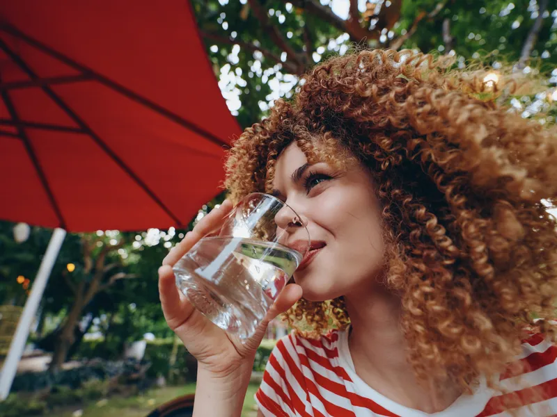 Close-up of a woman with curly hair sipping water from a glass outdoors, beneath a red umbrella, wearing a striped shirt, capturing a fresh and refreshing moment in warm light.