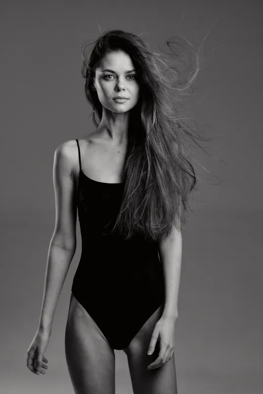 Black and white portrait of a young woman with long hair wearing a black bodysuit confidently posing against a plain background.