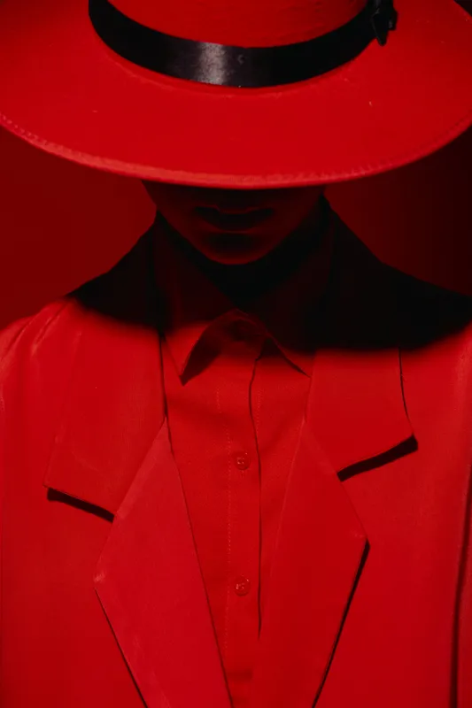 Figure in a red suit and wide-brimmed hat partially conceals the face showing minimal details. Dramatic lighting highlights textures of luxury fashion clothing. Creative concept for stylistic beauty.