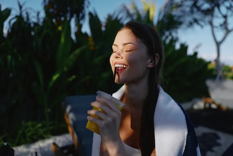 Young woman with towel wrapped around shoulders enjoys refreshing drink outdoors in sunny garden setting with closed eyes and relaxed happy expression on face