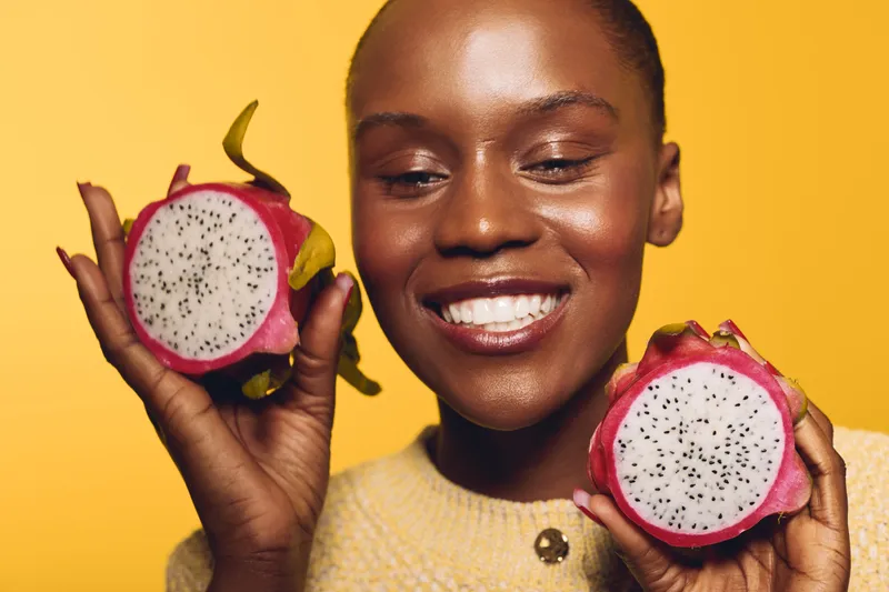 Smiling dark skinned African woman holds two halves of dragon fruit on yellow background. Concept of healthy lifestyle, exotic fruit, natural beauty, and nutrition for wellness.