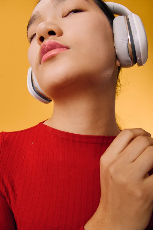 Woman wearing wireless headphones in red sweater listening to music with closed eyes, enjoying sound and relaxing on vibrant yellow background, closeup portrait of calm person.