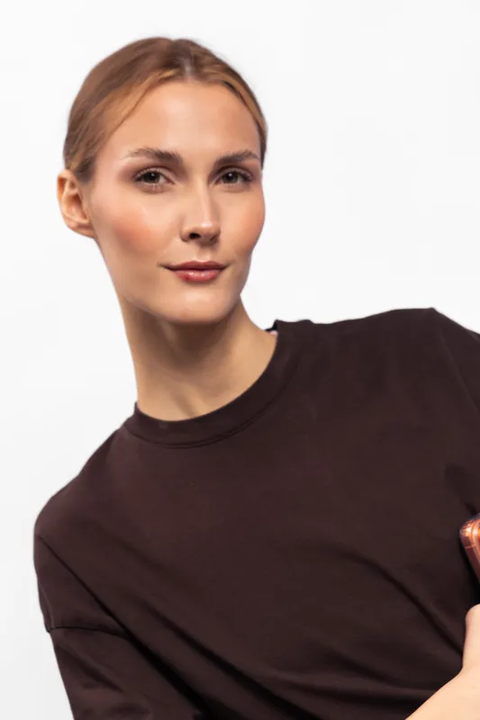 Confident young woman with fair skin and light brown hair wearing a dark brown shirt looks at camera with slight smile. Studio portrait on white background. Concept of beauty and confidence.