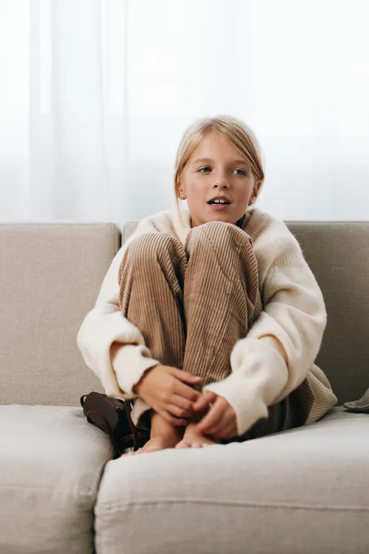 Authentic candid portrait of a young girl indoors, relaxed on a sofa in a cozy sweater, highlighting a simple school life moment and playful home scene.