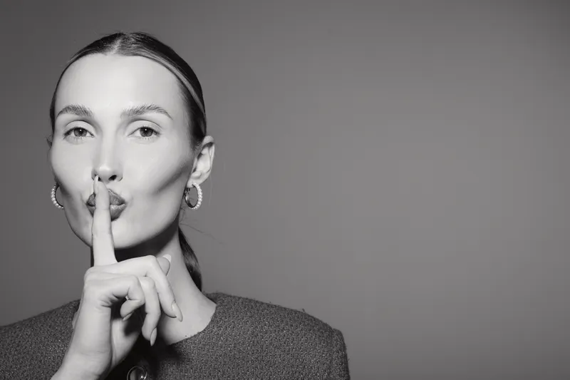 White woman with light skin and pulled back hair gestures silence with finger on lips against plain background. Concept for quiet, secrecy, and calm expression.