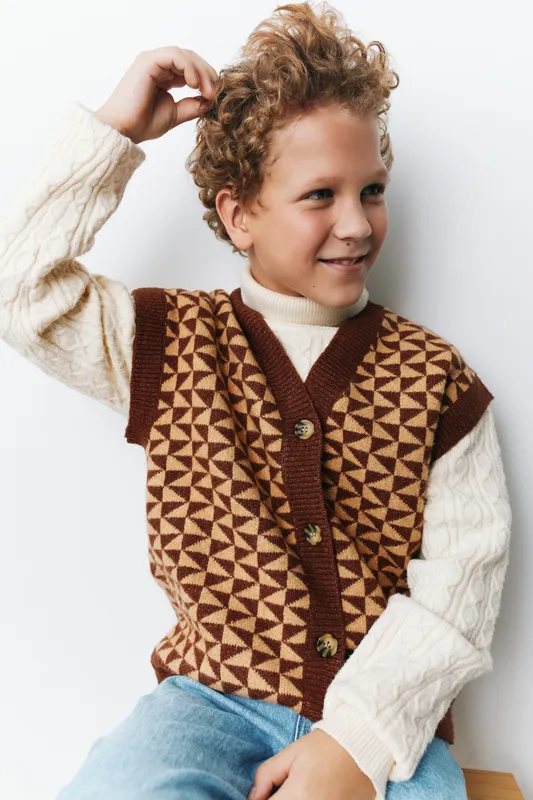 Portrait of a cheerful boy wearing a geometric knit vest over a cream sweater, seated against a light backdrop, capturing a candid moment and warm retro style.