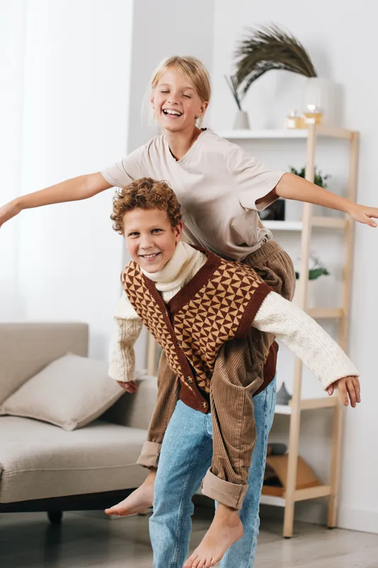 Two boys share a joyful piggyback ride indoors, capturing playful laughter and bright energy as they bounce around a cozy living room, creating a candid family moment.