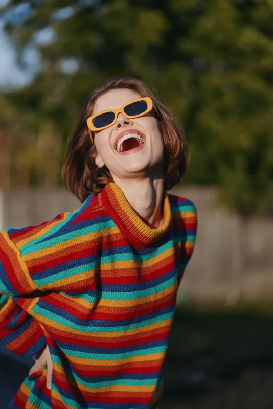 Woman laughing young adult in a colorful striped sweater and orange sunglasses, casual outfit outdoors. Joyful portrait with bob haircut, playful pose and vibrant lifestyle fashion vibe in sunlight.