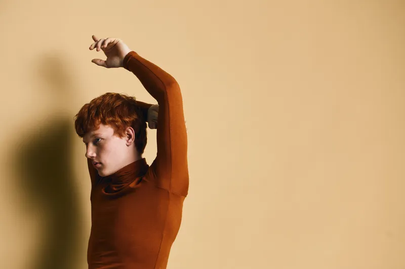 Portrait of young woman with red hair wearing brown turtleneck posing with raised arms against beige background for fashion and style concept.