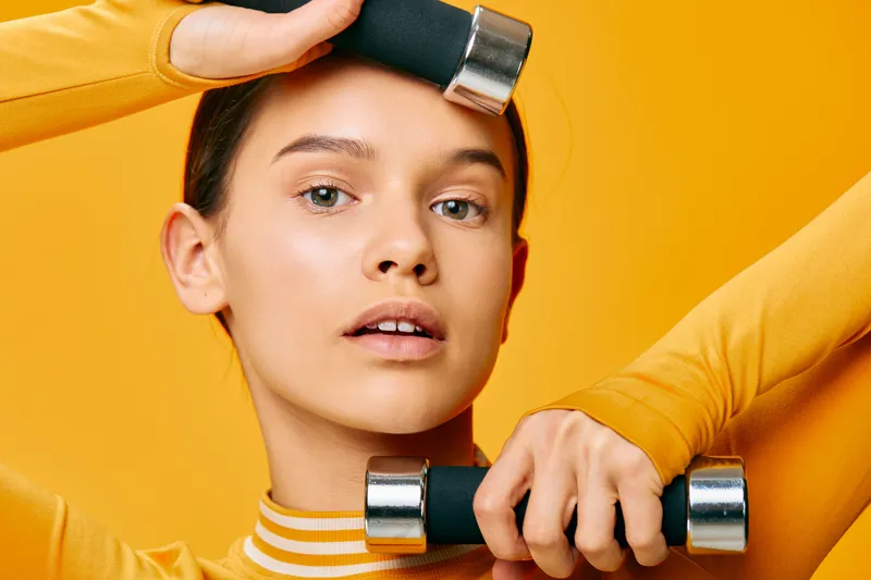 Woman in bright orange sportswear lifting dumbbells against a bold yellow background, focused expression, athletic workout scene emphasizing strength, balance and determination in fitness training.