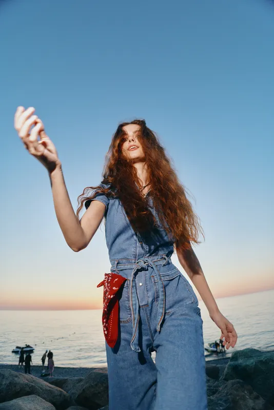 lifestyle woman in modern boho-western denim street style posing outdoors with film color effect during golden hour by the seaside, casual and stylish vibe