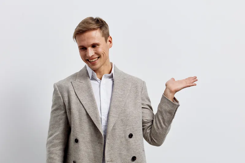 Confident young Caucasian man smiling, wearing a stylish gray blazer, posing with one hand raised, isolated on a plain white background. Studio portrait of a cheerful professional feeling positive and