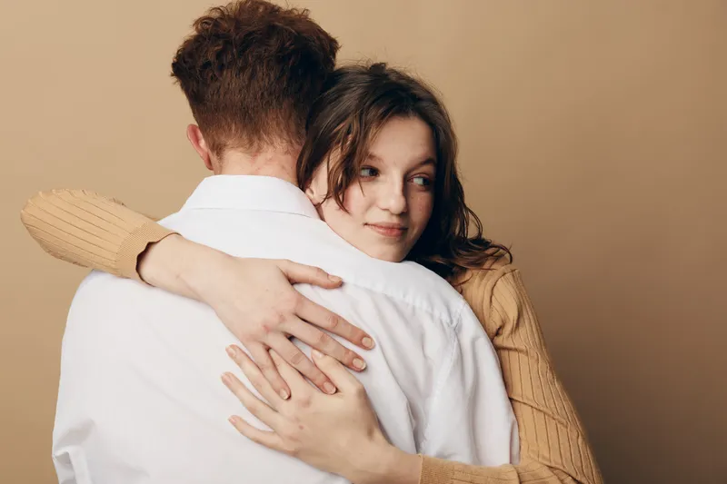 Young woman in beige sweater hugs man in white shirt from behind, showing affection and comfort. Warm neutral background creates intimate and tender mood of love and support.