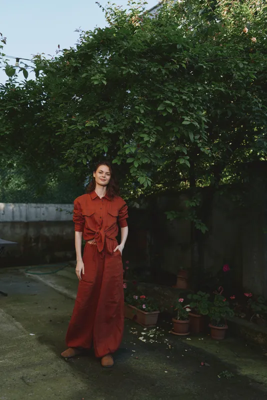 A woman in a red dress stands in a garden, posing with a relaxed posture. Bright daylight highlights lush greenery and potted plants. Focus on everyday fashion, outdoor living, and feminine