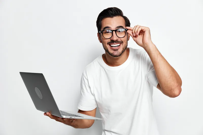 Smiling young man in casual white t-shirt adjusts glasses while holding laptop, looking at the camera with a cheerful expression. Technology and lifestyle concept.