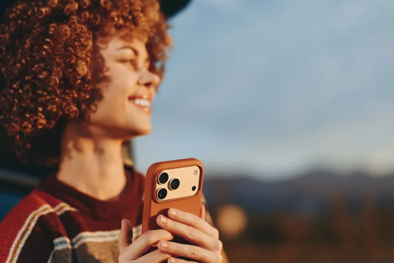Woman lifestyle outdoor portrait, curly hair, smiling and holding smartphone, side view profile, warm sunset light, blurred landscape background, casual everyday closeup moment