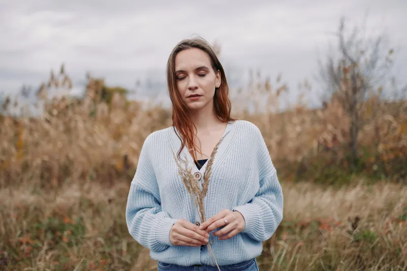 A contemplative young woman in a soft blue sweater gently holds dried meadow grass, captured in an authentic, natural moment that conveys honesty, quiet credibility and a calm, reflective mood.