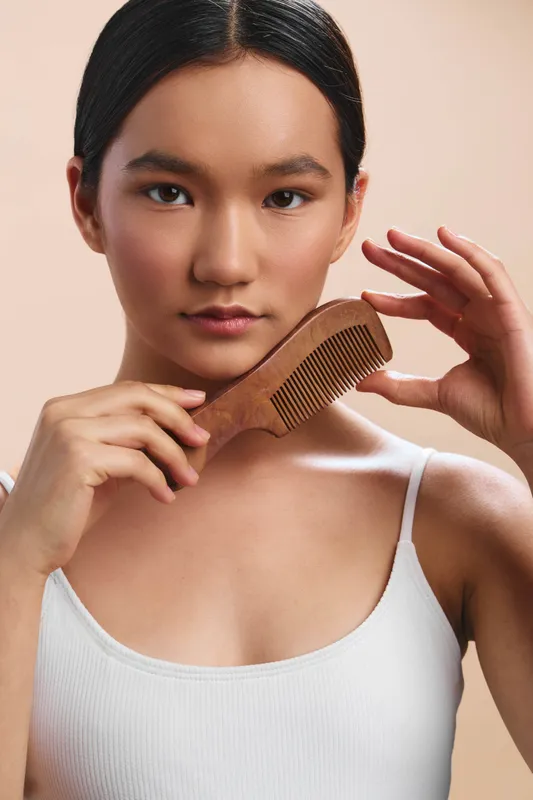 asian woman grooming hair with a wooden comb in a calm studio setting, focused smile, soft lighting, minimal background, everyday beauty routine, portrait style, warm and inviting mood