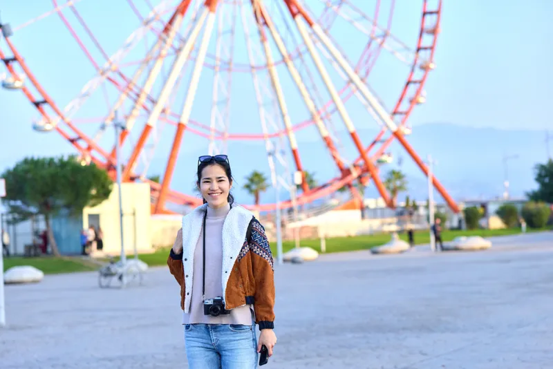 A woman with a camera at a sunny amusement park near a large ferris wheel, capturing joyful moments outdoors with casual fashion and vibrant rides in the background