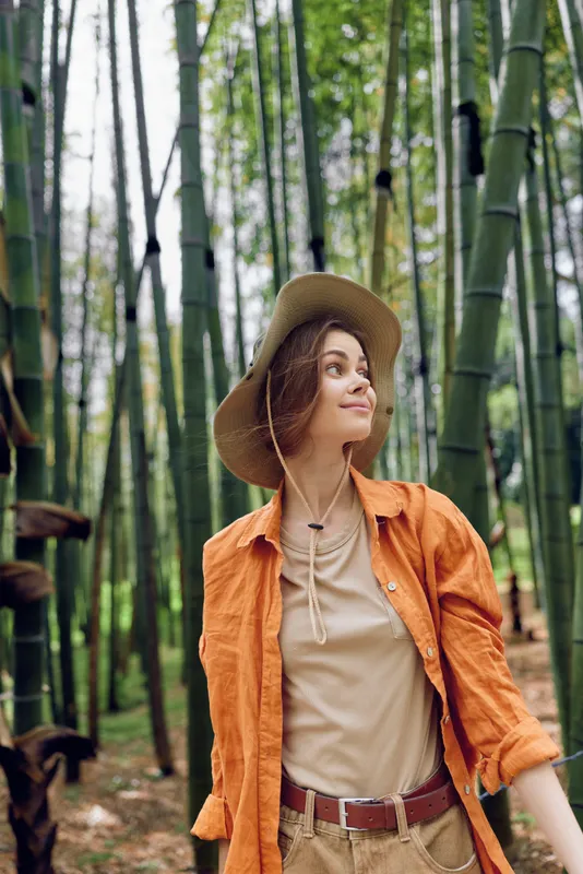 Woman hat portrait in a bamboo forest, looking up with a soft smile. Casual travel outfit with earthy jacket and relaxed nature mood for outdoor exploration and serene lifestyle.