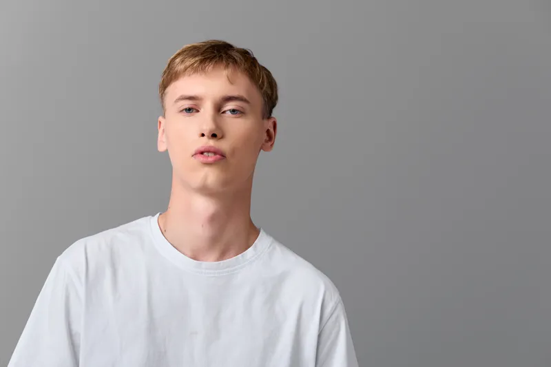 Young man in a white t shirt stands before a neutral background in a studio setup, with calm expression and soft lighting for a clean, fashion oriented image.