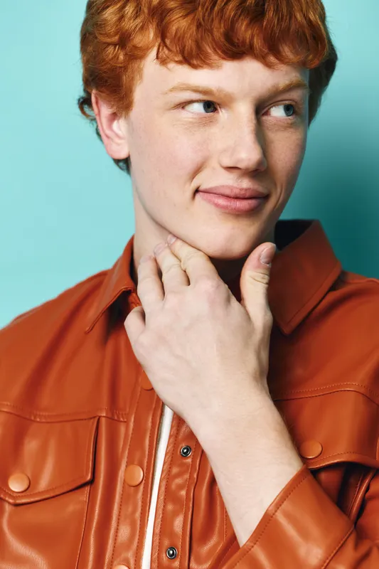 Redhead young man wearing a brown leather jacket touching his neck with thoughtful expression on blue background. Portrait of confident male with fair skin and short hair.