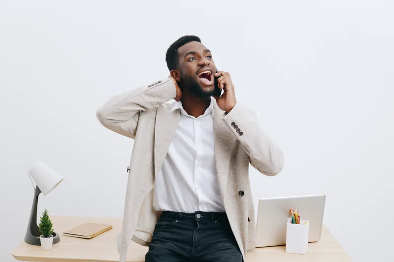Smiling man talking on the phone in bright modern office with laptop, desk, and supplies, expressing happiness and engagement during work or casual conversation, lifestyle concept, casual professional