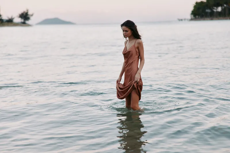 A lone woman stands in calm shoreline water at dusk, wearing a flowing dress that grazes the surface. Gentle waves, soft light, and tranquil seaside mood.