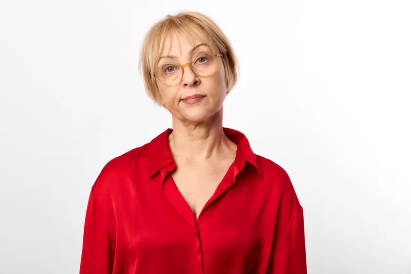 Senior woman portrait with glasses and a red shirt, neutral calm expression against a white studio background, soft light headshot for wellbeing and confidence